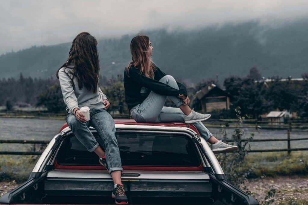 Amigas sentadas em carro, paisagem montanhosa ao fundo.