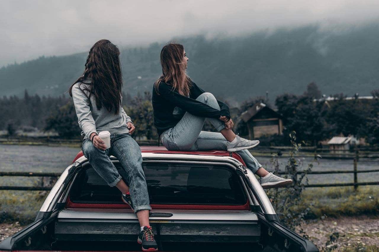 Amigas sentadas em carro, paisagem montanhosa ao fundo.