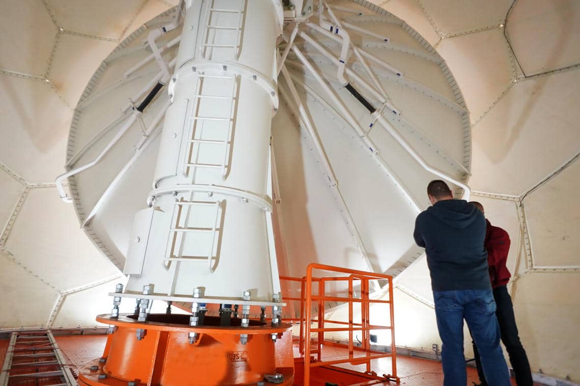 Pessoas observam equipamento grande em estrutura metálica.