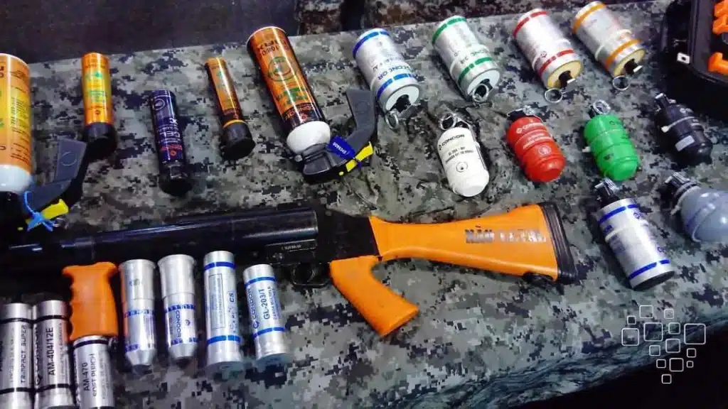 Armas não letais e cartuchos sobre mesa camuflada.