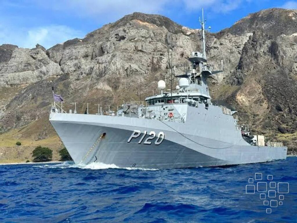 Navio de guerra navegando em mar azul, paisagem rochosa.